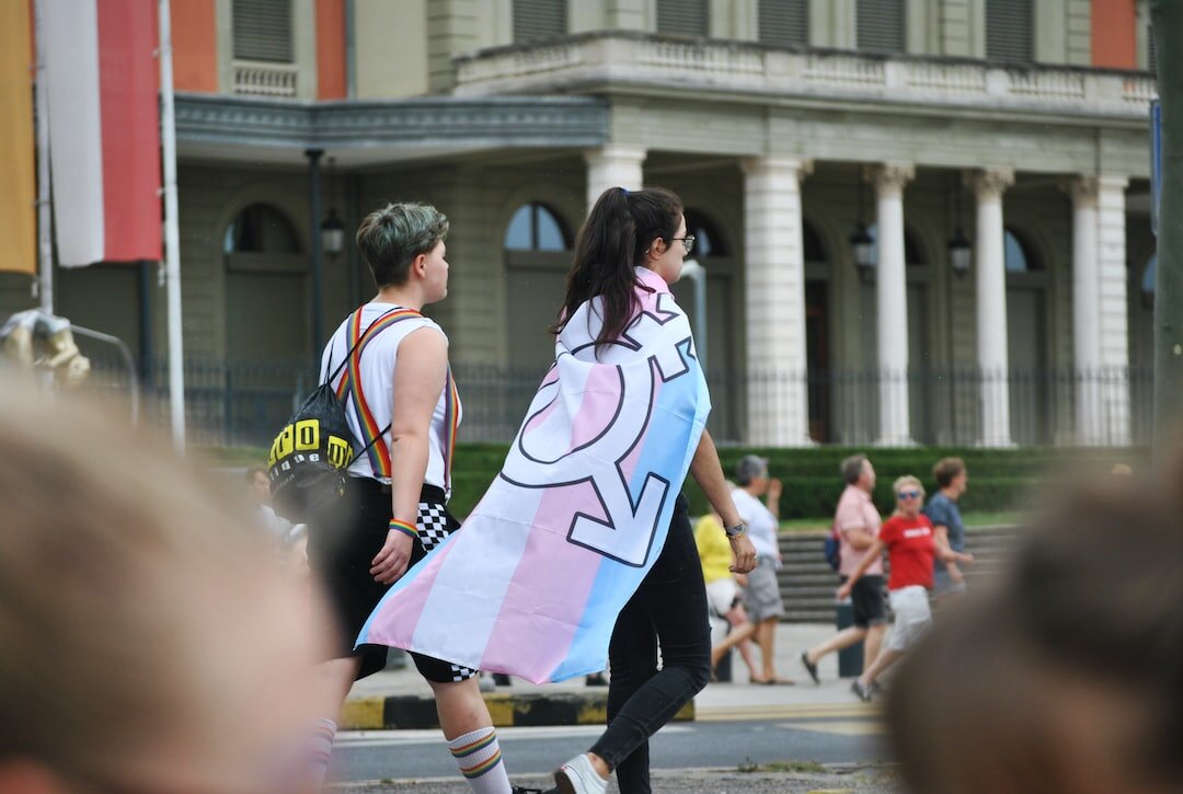 Two people in LGBTQ+ pride parade one with transgender pride cape